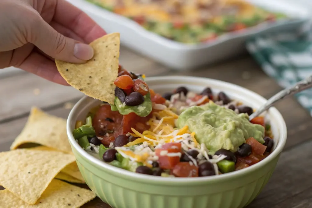 A hand scooping into a colorful layered dip with a tortilla chip